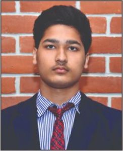 Portrait of a young man in a dark suit with a red tie, standing in front of a brick wall.