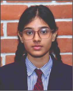 Young girl with glasses in a school uniform (blazer, striped shirt, red tie) standing against a brick wall with braids.