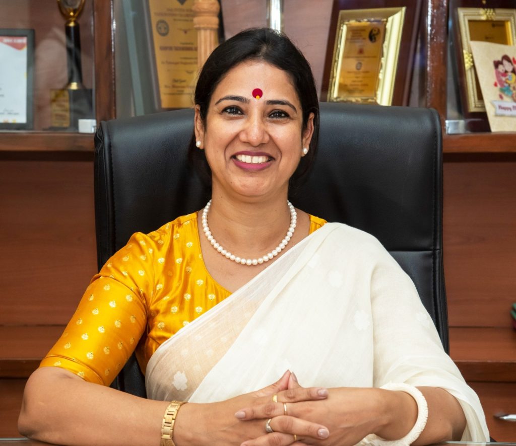 Smiling Indian woman in a yellow blouse and white sari sits at a desk in an office, wearing pearl necklace and gold jewelry, with awards on the shelf behind her.