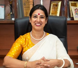 Smiling Indian woman in a yellow blouse and white sari sits at a desk in an office, wearing pearl necklace and gold jewelry, with awards on the shelf behind her.
