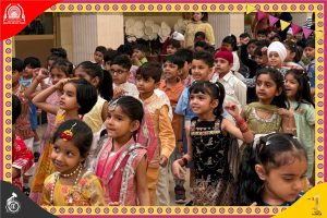 A group of young Indian children in colorful traditional clothes dancing together at a school event. In festive outfits, they smile and move in a coordinated line in a courtyard setting.