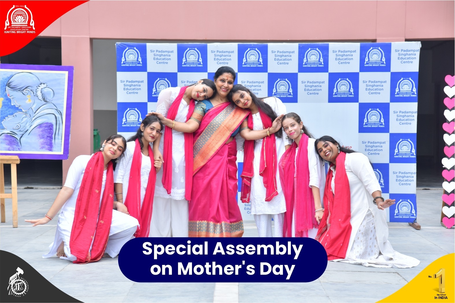 Group of women in white outfits with pink scarves posing together for a Mother's Day event, in front of a school backdrop and banner saying 'Special Assembly on Mother's Day'