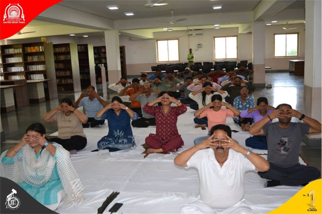 Group of adults sitting on a white mat in a library-like room doing eye exercises with hands over their eyes.