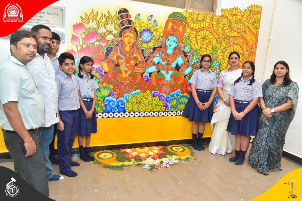 Group of students in uniform posing with two teachers beside a bright mural of Hindu deities Krishna and Radha, with a flower rangoli on the floor.