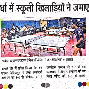 Table tennis match in a hall with players at a long table and spectators watching from the sides.