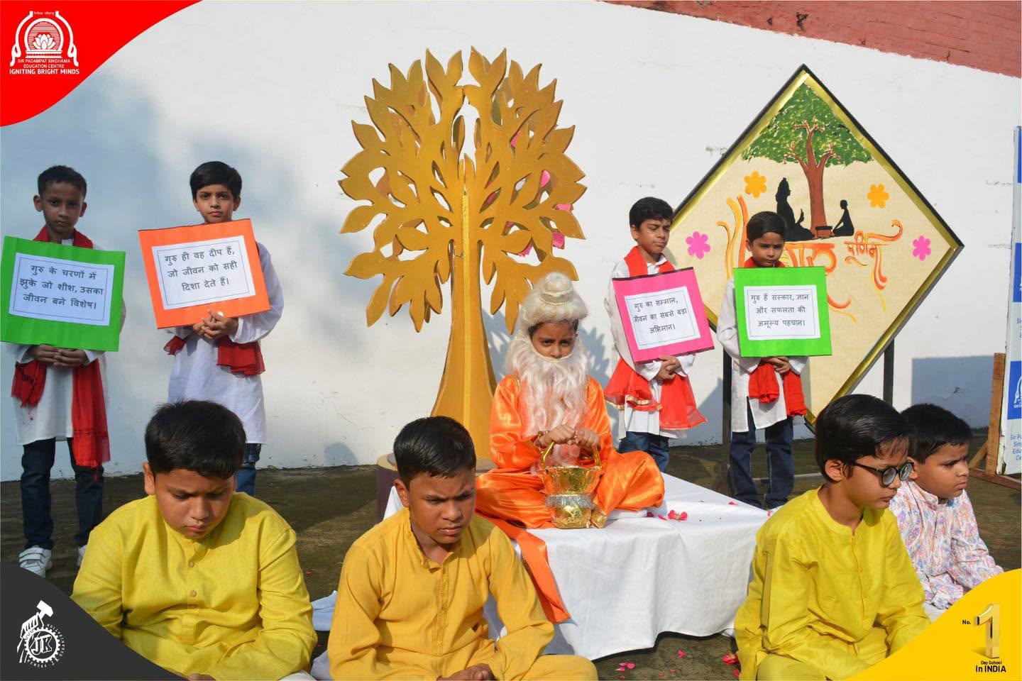 School event scene with kids in traditional outfits, a bearded man in orange robes seated on a white cloth, and students holding colorful message signs behind a golden tree backdrop.
