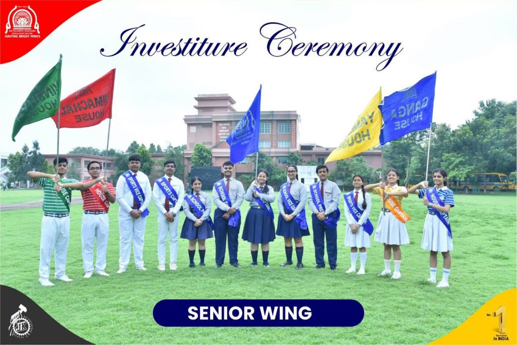 Students in uniform with blue sashes stand on a grassy field during an investiture ceremony; a bold 'Senior Wing' banner sits at the bottom.