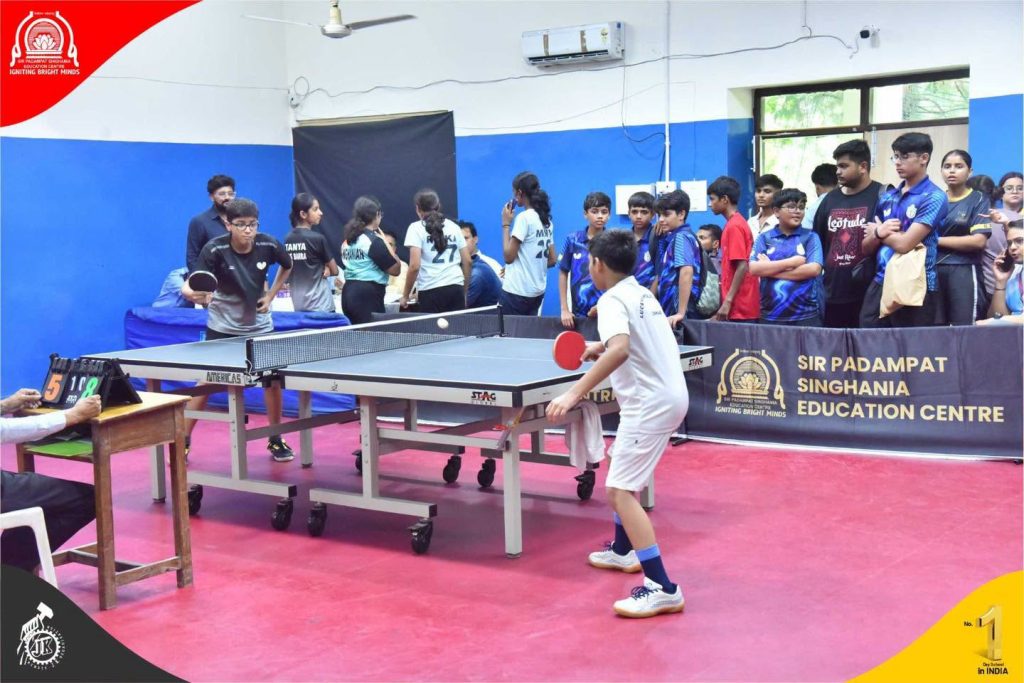 SPSEC Hosts CBSE Cluster-IV Table Tennis Championship 2026 Two boys play a table tennis match in a school gym, with a referee at a side table and a crowd of students watching behind a barrier with a school banner.