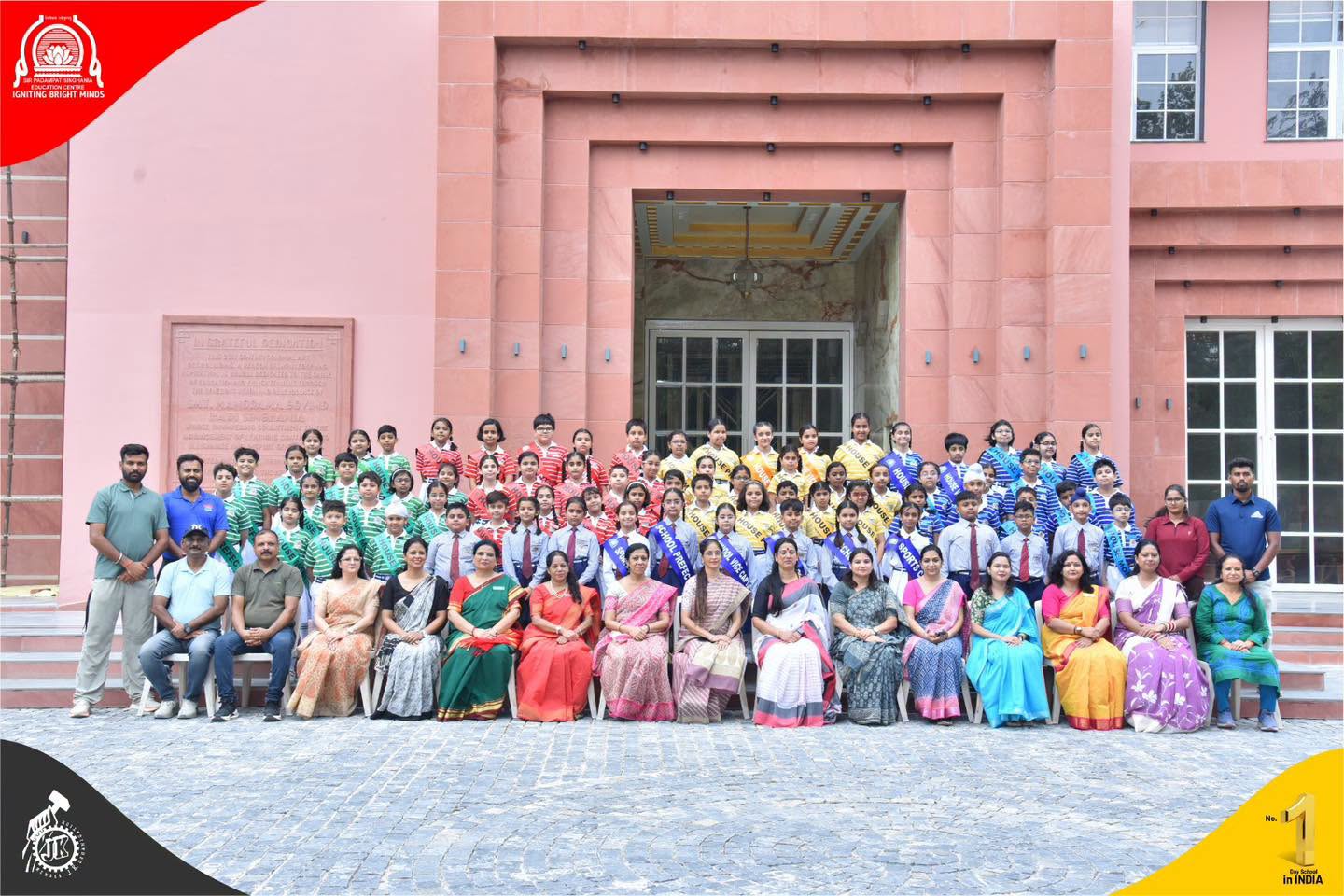Group photo of students and teachers outside a pink school building, arranged in rows by uniform colors on a cobblestone courtyard.